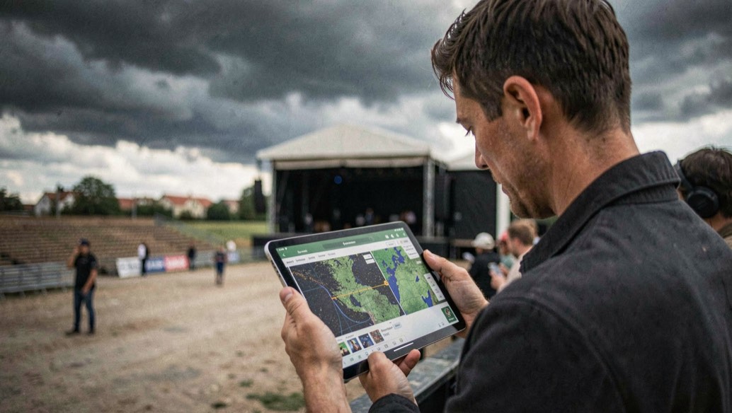 Event crew studies weather radar on tablet beneath darkening clouds near empty stage.