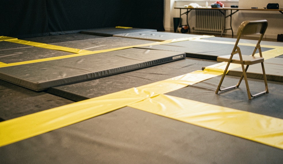 A stunt rehearsal floor with thick mats, bright blocking tape and a folding chair under warm studio light.