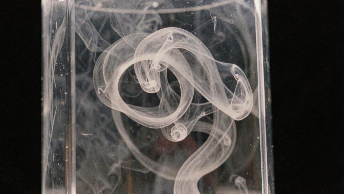Smoke ring curls through transparent chamber lit against black background.